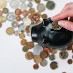 Close-up of a hand inserting a coin into a black piggy bank with scattered coins on a white background.