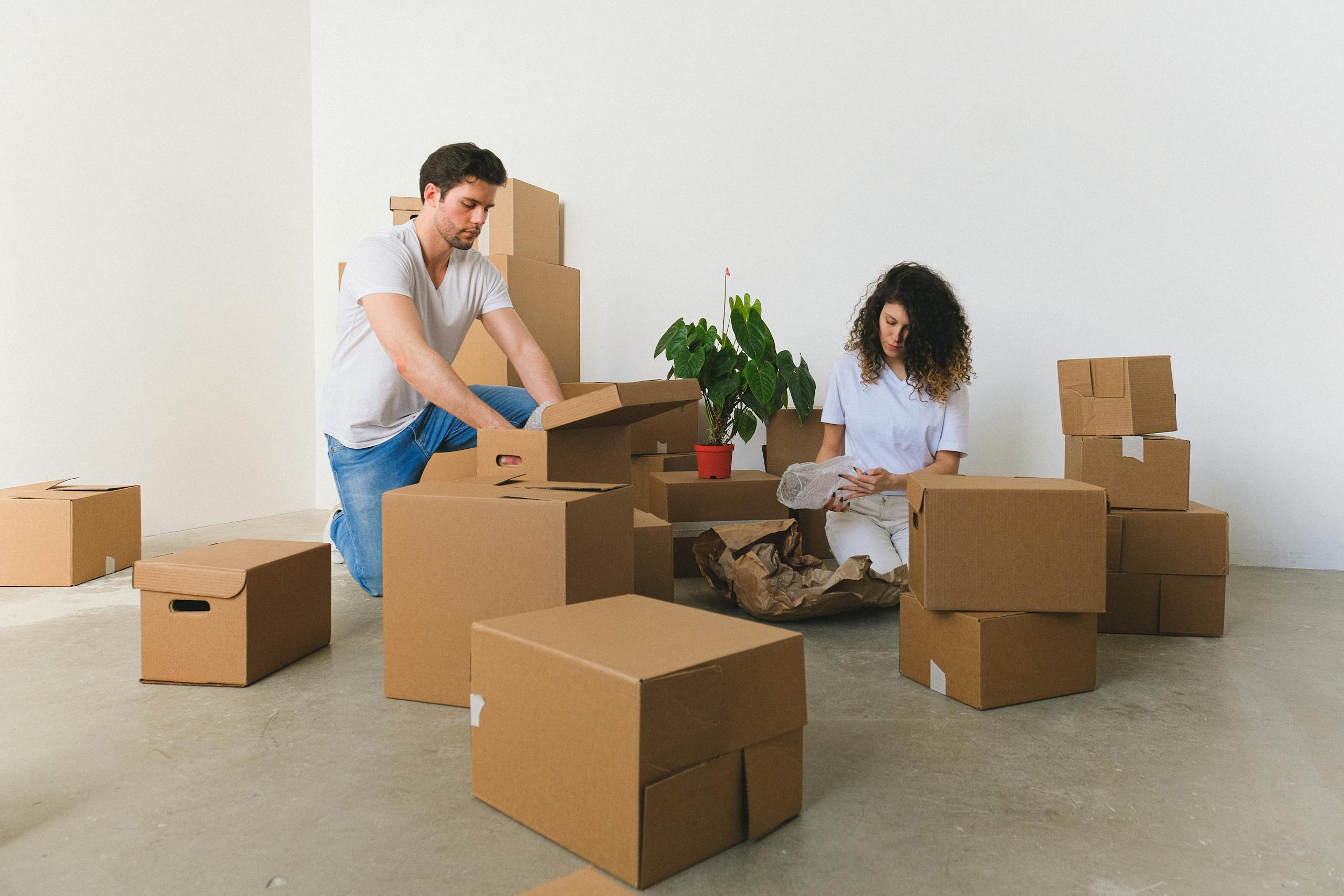 Serious couple opening brown large carton boxes with different belongings on floor at home